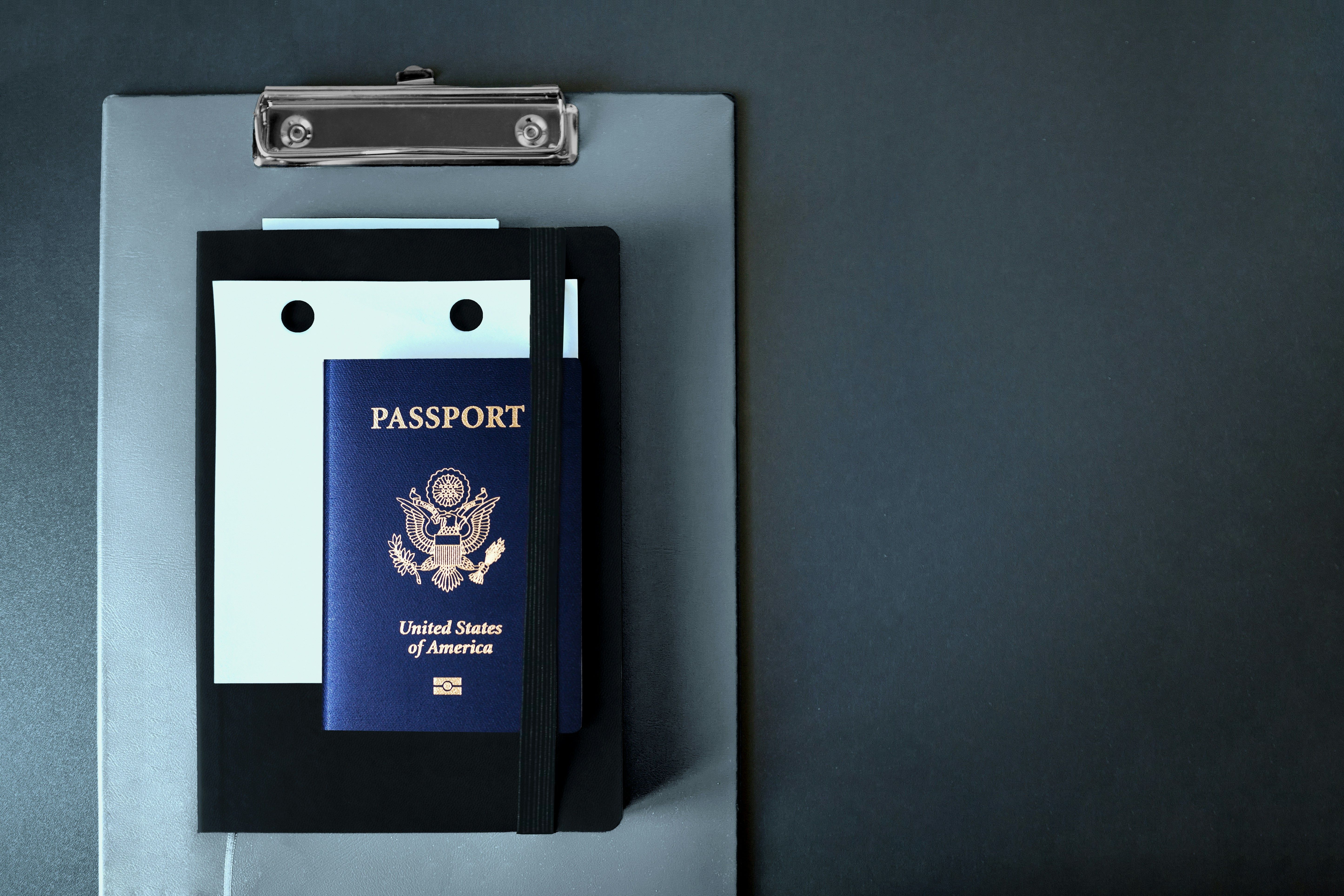 A dark blue United States passport lies on top of a clipboard with white papers.
