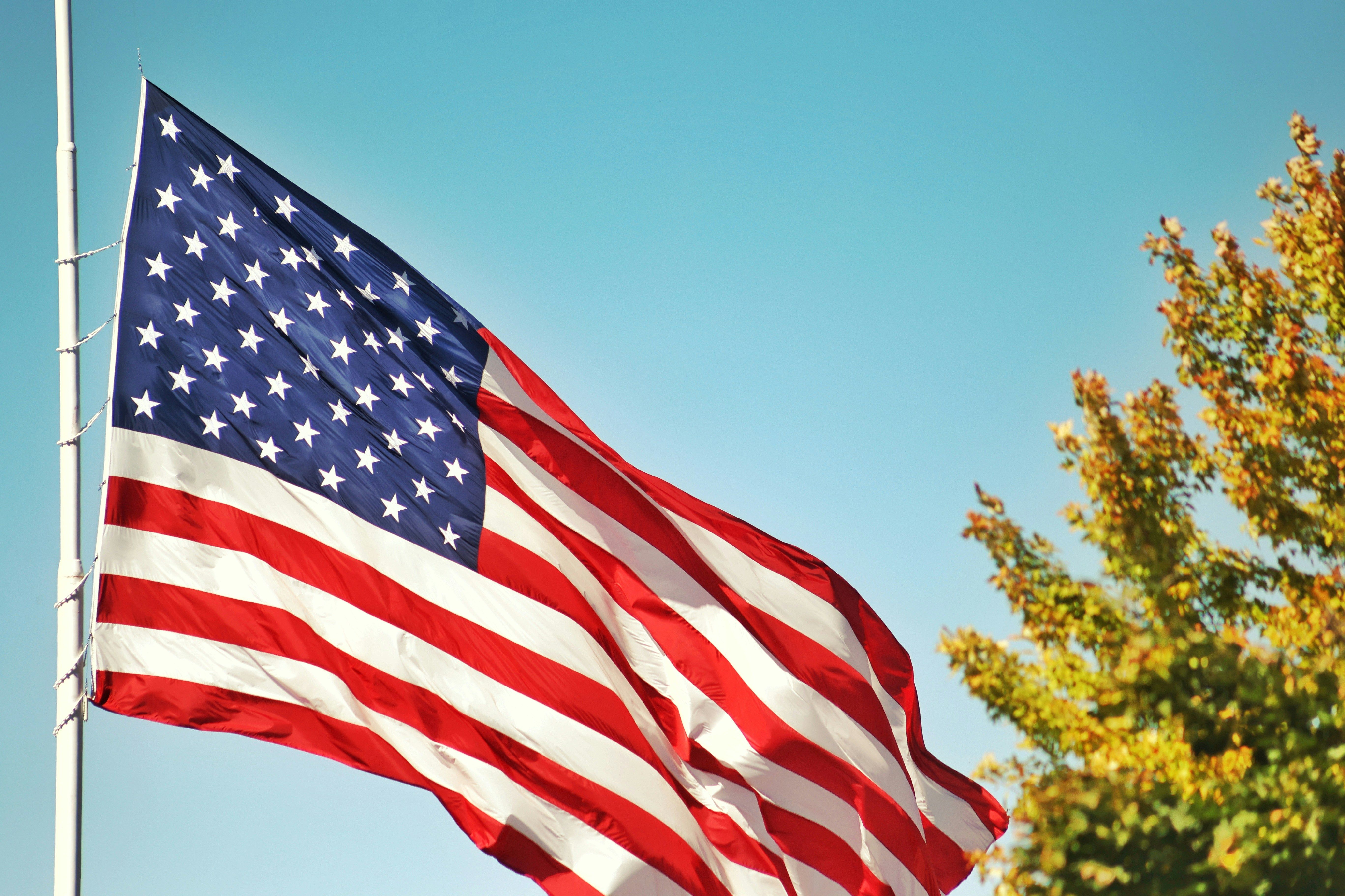 American flag flying at half-mast against a clear blue sky with autumn tree leaves.